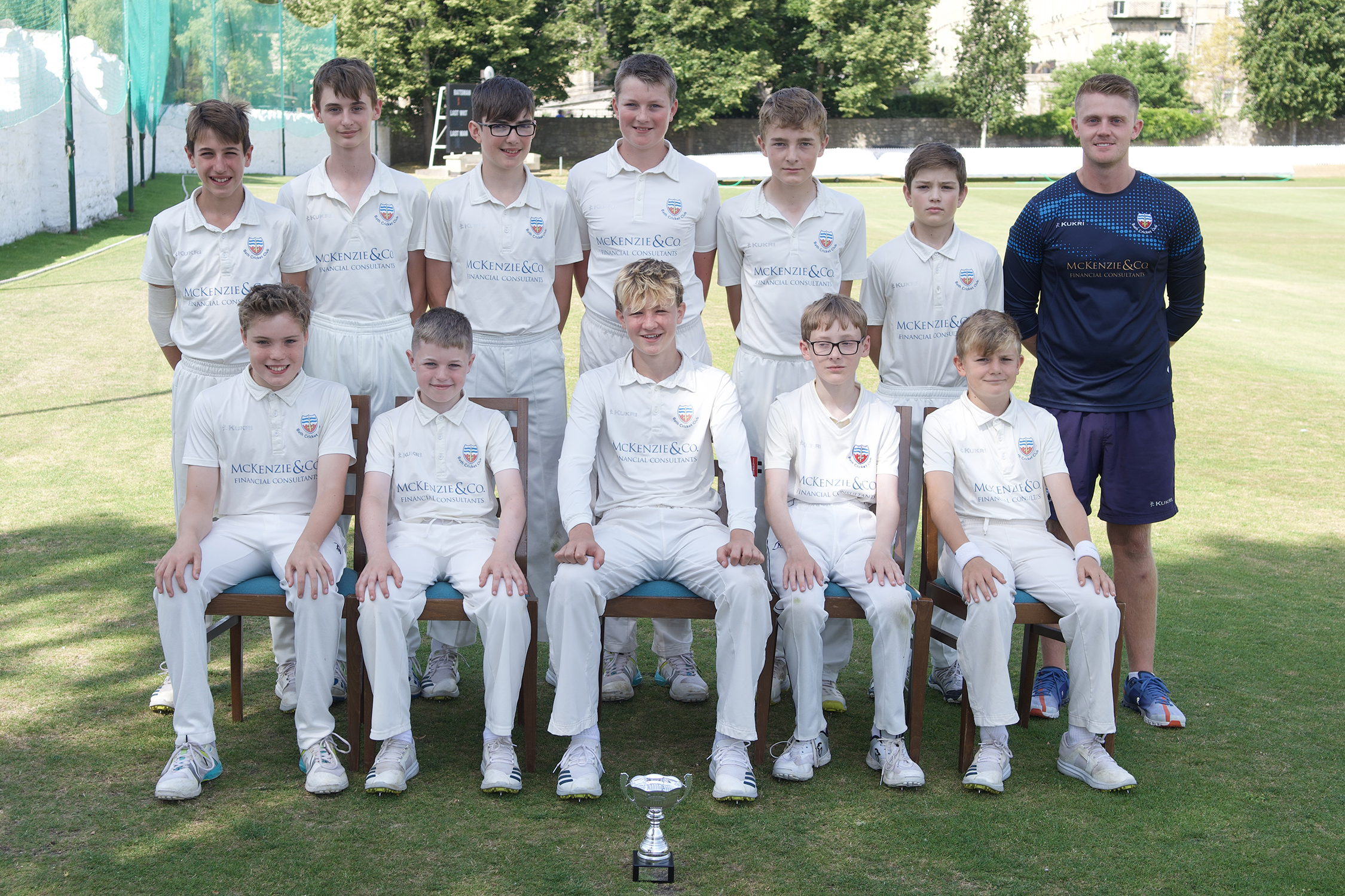 Team photo of 11 players and one coach taken on a sunny day with cricket ground behind